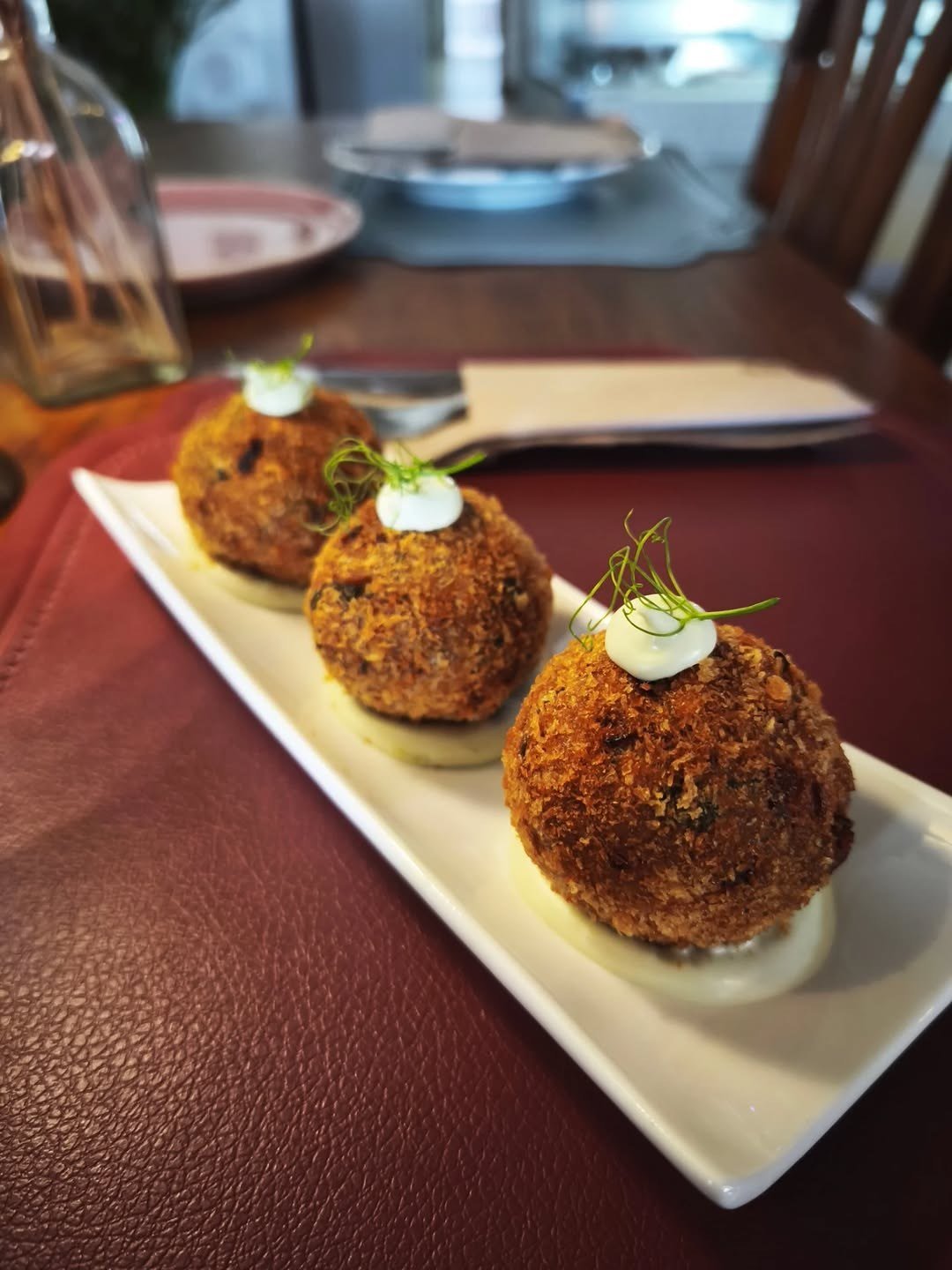 Três croquetes panados numa travessa branca, cada um com uma colher de maionese e ervas por cima