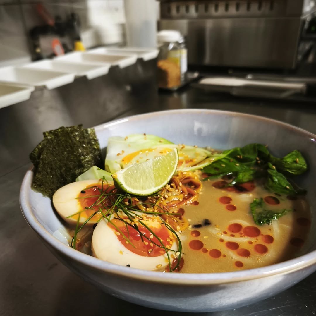 Taça de ramen com ovos cortados ao meio, alga nori, rodela de lima e ervas frescas