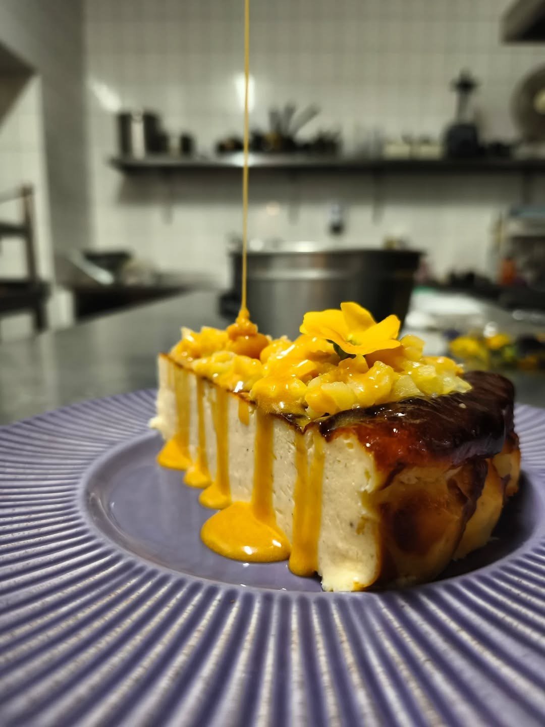 Fatia de cheesecake com caramelo a escorrer, num prato azul, com a cozinha de inox ao fundo